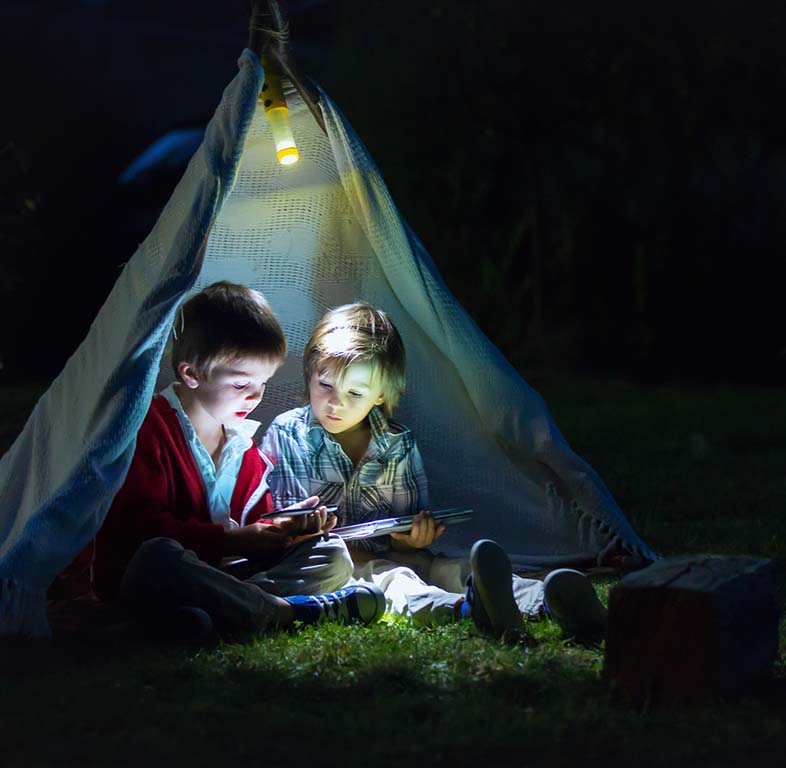 Cute little brothers, playing on tablet and telephone at night in camp side, in the tent