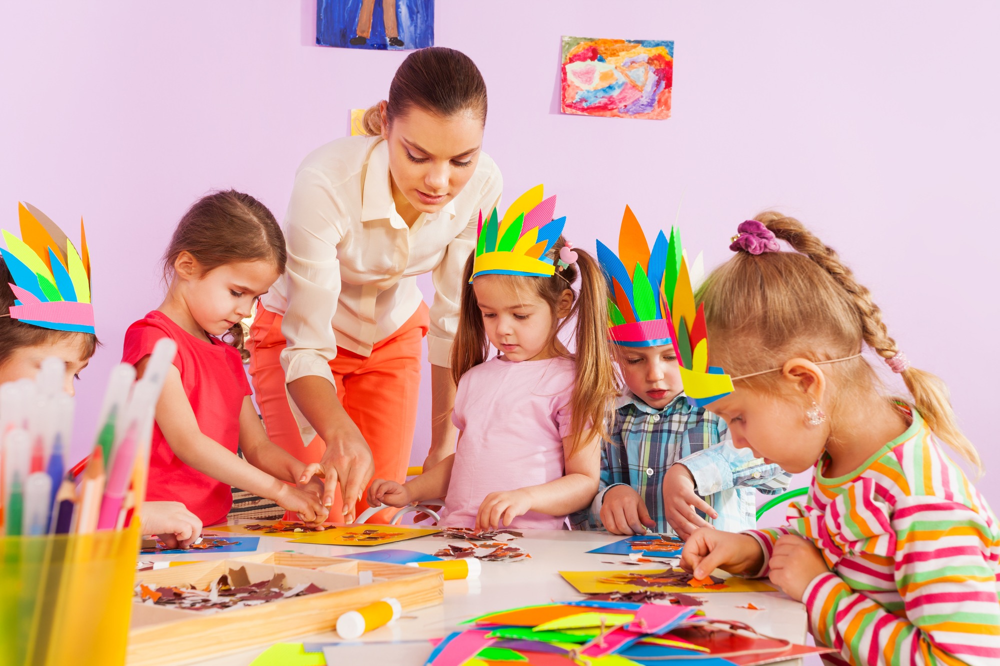 A teacher is guiding a group of young children wearing colorful paper crowns as they work together on arts and crafts at a table