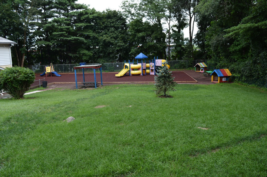 A spacious outdoor playground with colorful equipment including slides, climbing structures, and playhouses, set on brown safety surfacing with a green lawn in the foreground and trees in the background