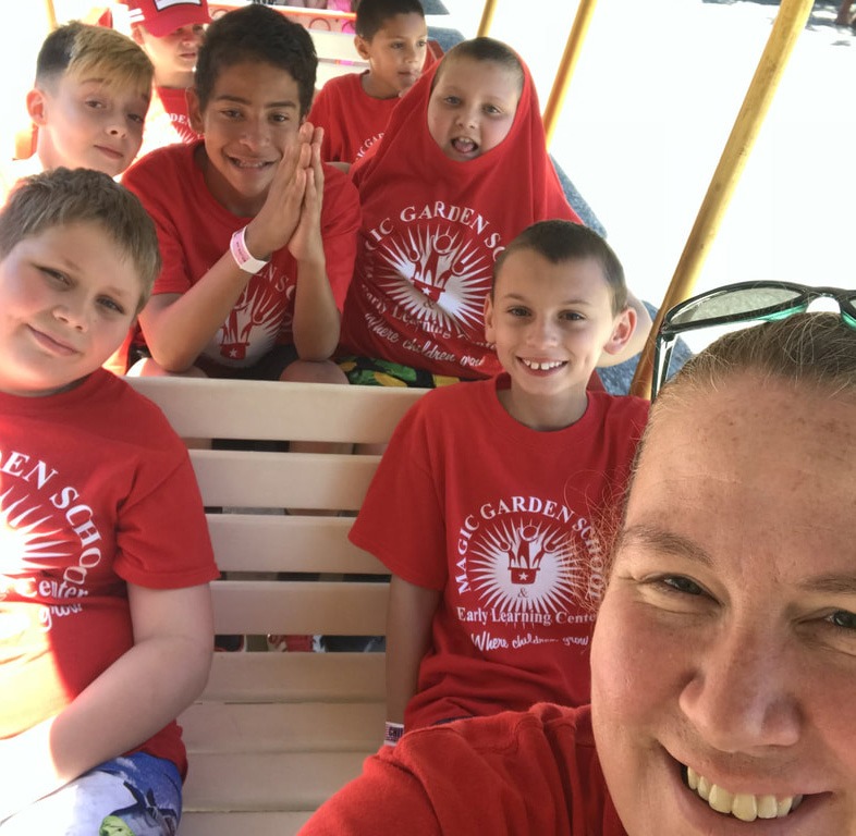 A group selfie of smiling children and an adult wearing matching red "Magic Garden" t-shirts, appearing to be on a field trip or group outing