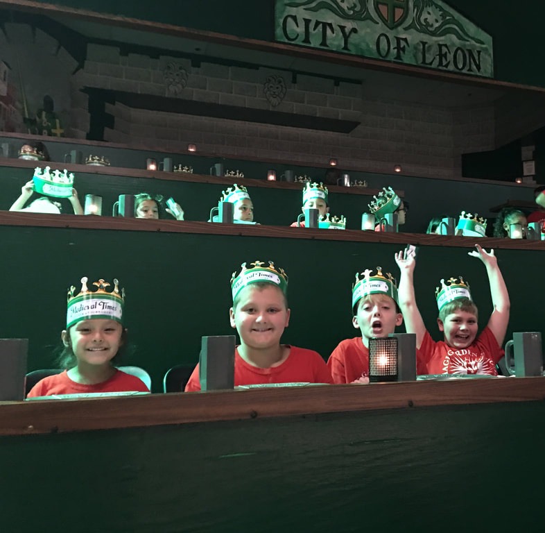 Four children wearing paper crowns and red shirts sit excitedly in theater-style seating at what appears to be a Medieval Times-style venue called "City of Leon."
