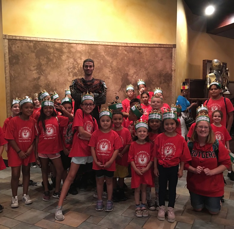 A group of children wearing matching red t-shirts and paper crowns poses with a tall man in medieval costume for a group photo indoors