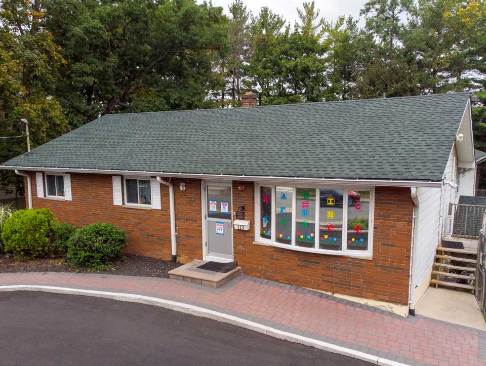 A single-story brick building with colorful children's artwork displayed in the windows, featuring a green shingled roof and surrounded by trees