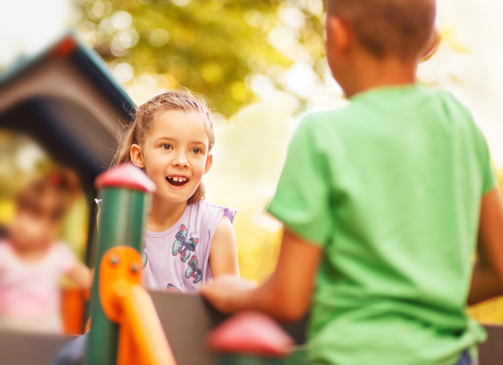 A young girl with an excited expression plays on playground equipment alongside another child wearing a green shirt
