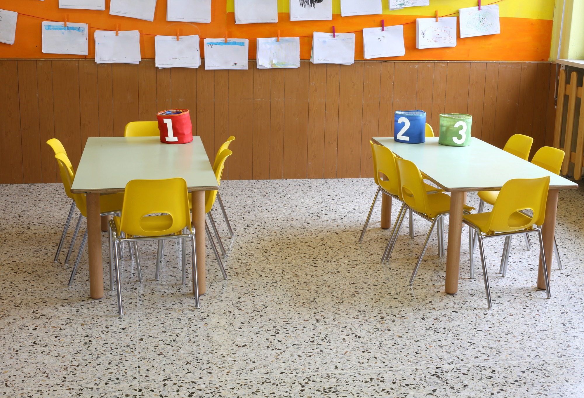 A bright preschool classroom with two white tables surrounded by yellow chairs, numbered storage containers (1, 2, 3), and children's artwork hanging on clotheslines above wood-paneled walls with orange and yellow accents