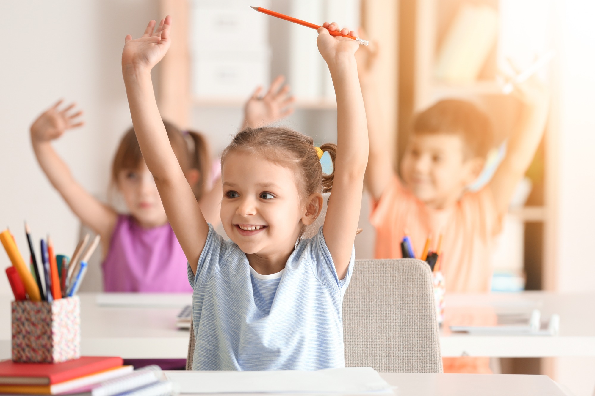 A young girl joyfully raises her hands with a pencil in a bright classroom, with other children raising their hands in the background