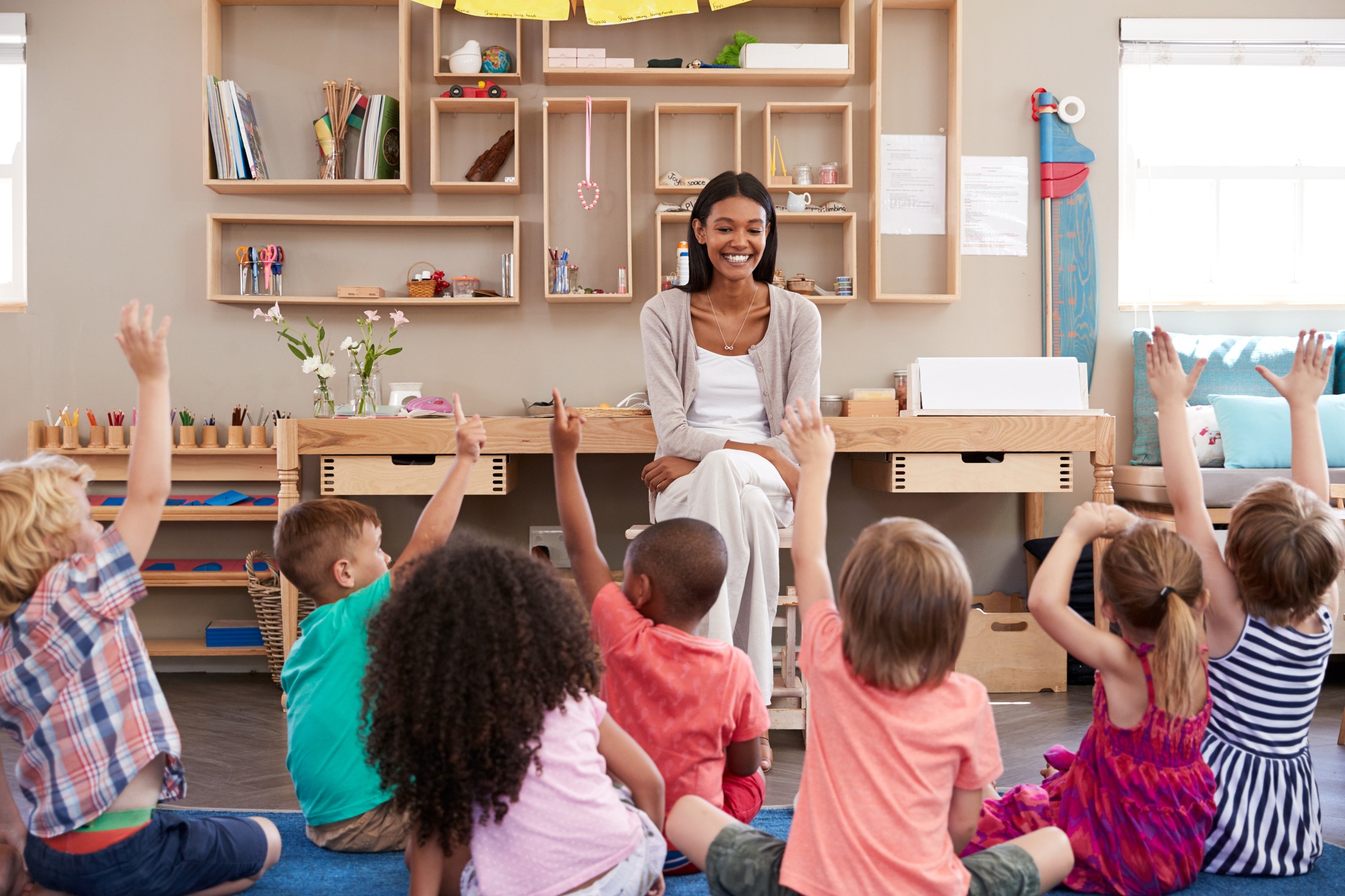 An enthusiastic teacher sits at a desk in a bright Montessori-style classroom while diverse children seated on the floor eagerly raise their hands to participate